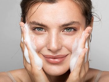 A young woman smiling while cleansing her face with a rich, white foam, demonstrating effective face wash or cleanser application for fresh, clean skin.