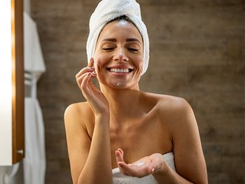 A happy woman with a towel wrapped around her head, applying a white cream or moisturizer to her smiling face, illustrating a post-shower skincare ritual or hydrating routine.