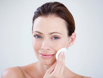 A beautiful woman with blue eyes gently wiping her cheek with a white cotton pad, suggesting makeup removal, toner application, or cleansing in her daily skincare routine.