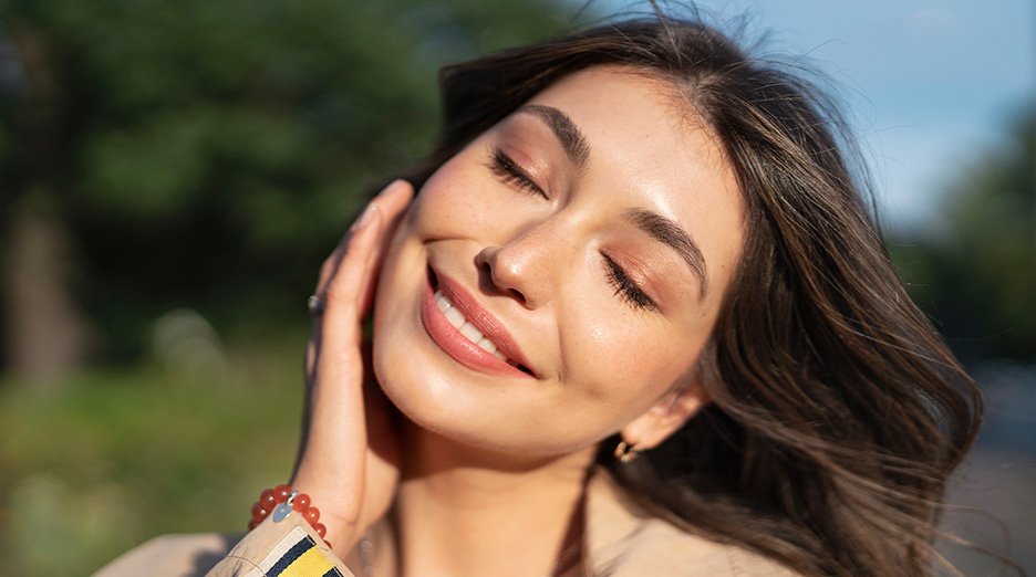 Front shot of a woman smiling with her eyes closed. One hand softly placed on her cheek as she tilts her head to take in the sun’s light and warmth.