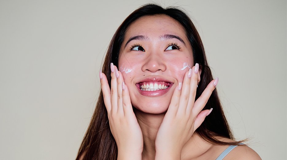 Front shot of woman smiling looking up as she applies moisturizer to both cheeks with both of her hands.