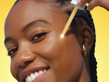 Brown skin model applying vitamin c serum to side of her cheek as she smiles at the camera.