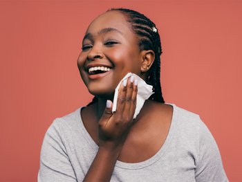 A young woman with dark skin smiles as she wipes her face with a cotton round containing an exfoliating toner.