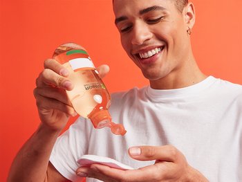 A young man with smiles as he pours toner onto a cotton pad, ready to apply it to his face.