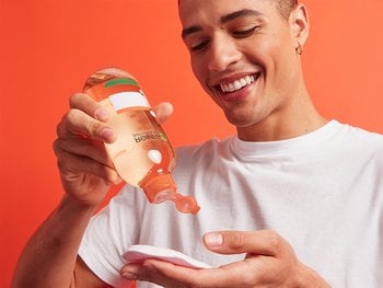 A young man with smiles as he pours toner onto a cotton pad, ready to apply it to his face.