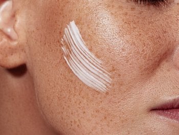 A close-up shows moisturizer being applied to a cheek and jawline area with fair skin.