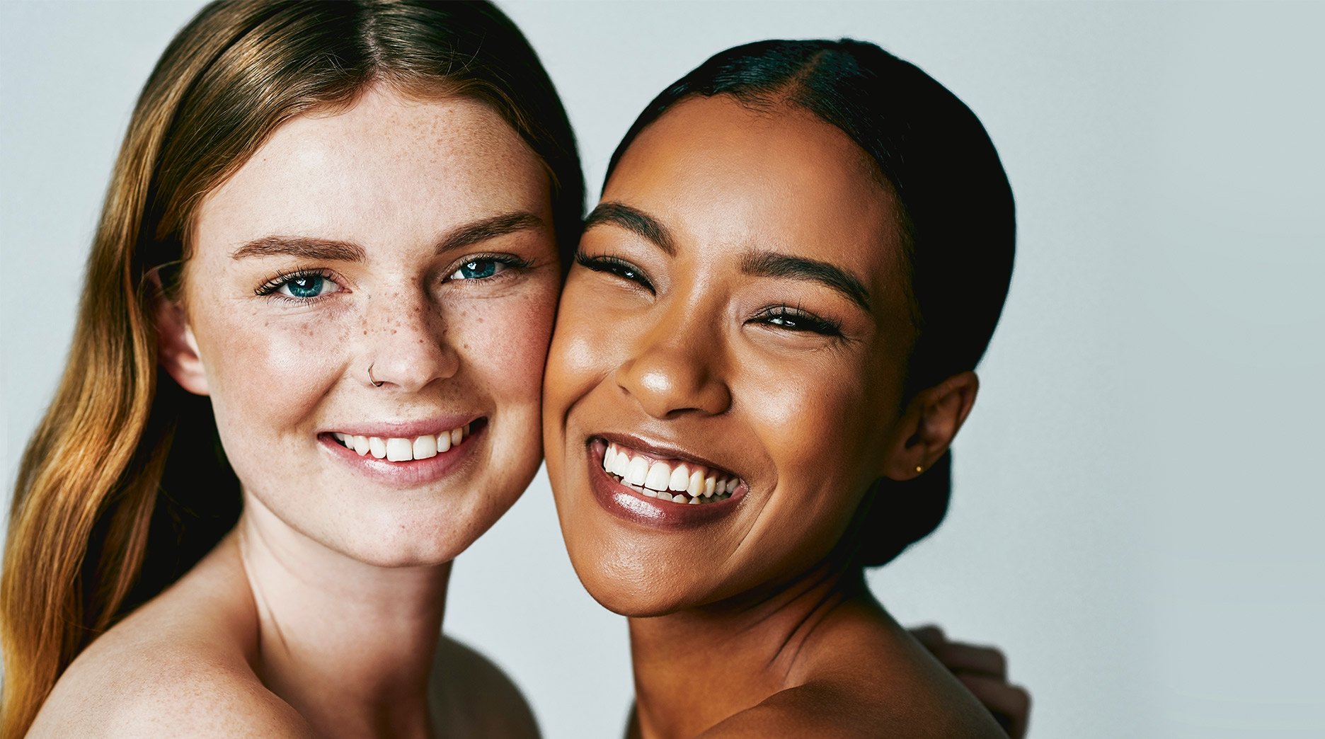 Two teenage girls, one light-skinned with freckles and red hair, the other dark-skinned with dark hair, smile with their cheeks touching, conveying a sense of youthful vibrancy and friendship.
