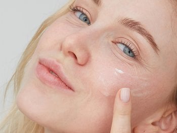 White, blonde female applying a light cream to her chees while smirking at the camera.