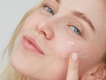 White, blonde female applying a light cream to her chees while smirking at the camera.