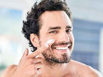 Front shot of man looking in the mirror, smiling as he applies face cream to his cheek.