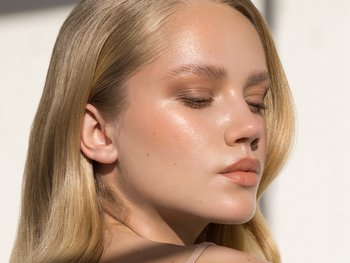 Front shot of blonde woman facing back as the sun makes her smooth skin glow. She has a clear, soft, glowing face.