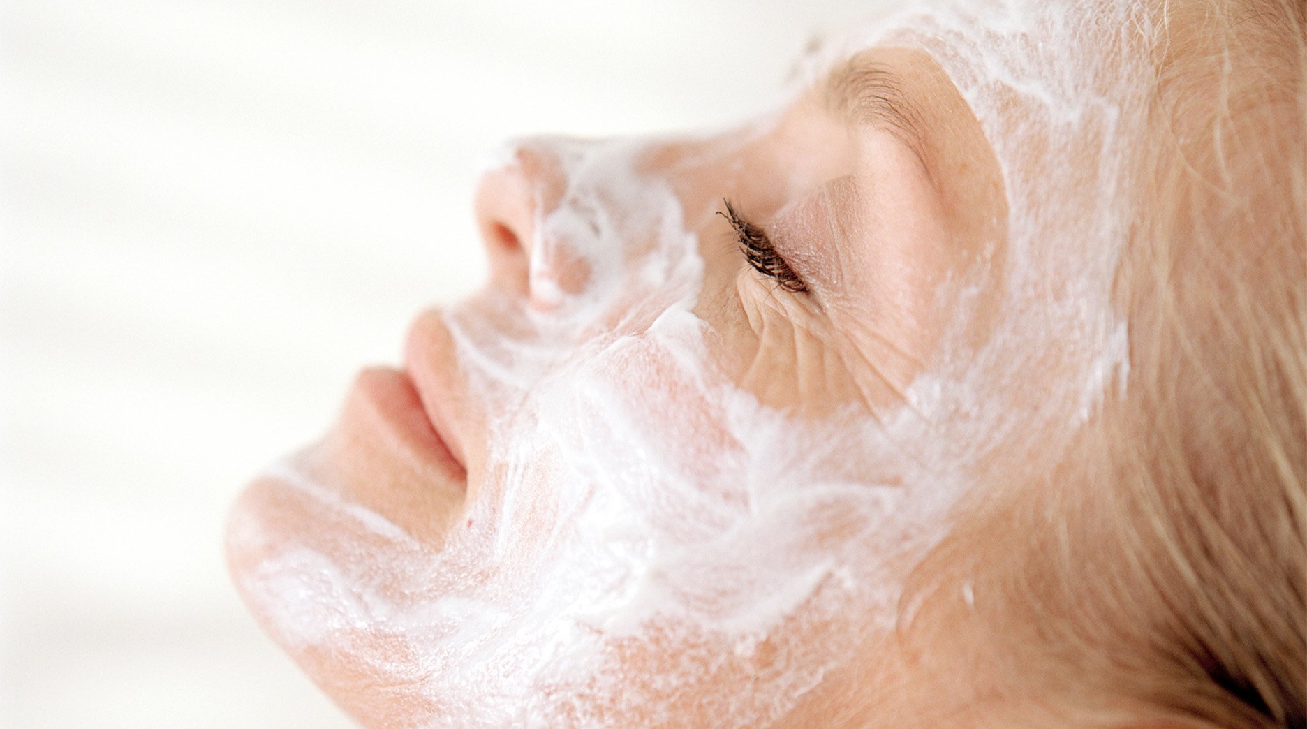 A side profile shot of an older woman relaxing with her head on a pillow as she lets her white clay mask rehydrate her skin. 