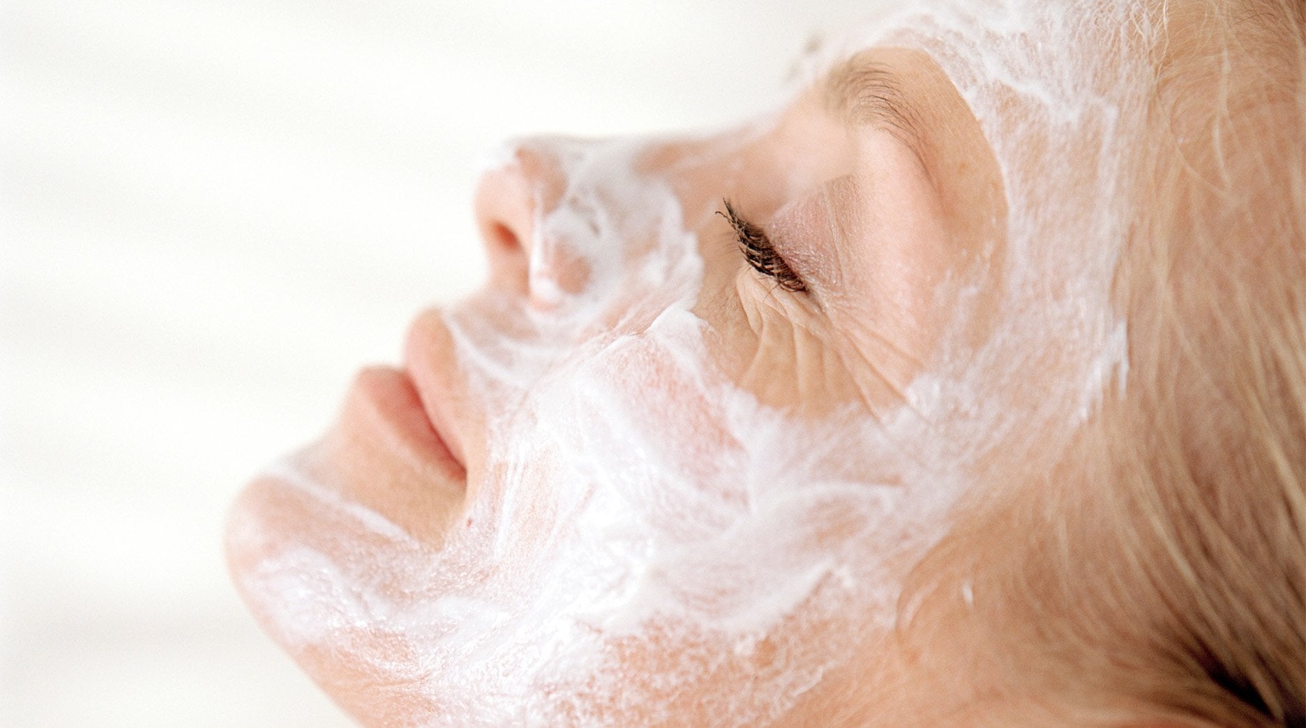 A side profile shot of an older woman relaxing with her head on a pillow as she lets her white clay mask rehydrate her skin. 
