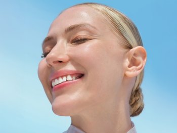 A smiling person with light skin and blonde hair looks up towards the sun.