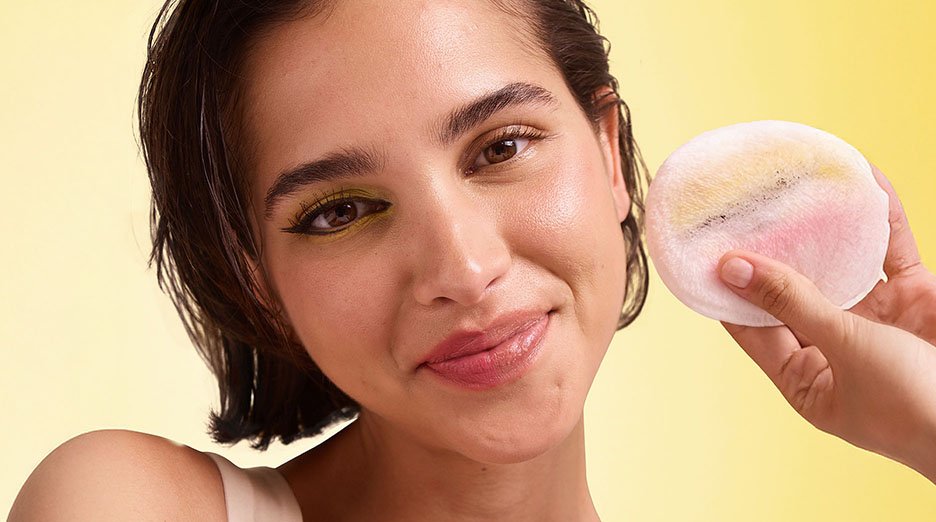 A woman with short, dark hair and expressive eye makeup (green and black eyeliner) holding a makeup remover pad with colorful makeup residue, smiling subtly at the camera against a yellow background.