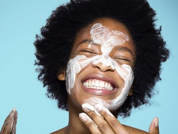 A woman with her hair pulled back smiles as she applies cleanser to her face, demonstrating the first step in any skincare routine.