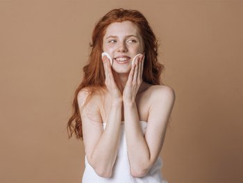 A woman with red hair applies toner to her face, illustrating the second step, toning, after cleansing.