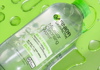 Top shot of Micellar Cleansing Water Mattifying, green cap, bottle. The bottle is lying horizontally on a green surface, on top of large water droplets.