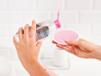 A hand pours micellar water onto a pink cotton round, showcasing its use and application.