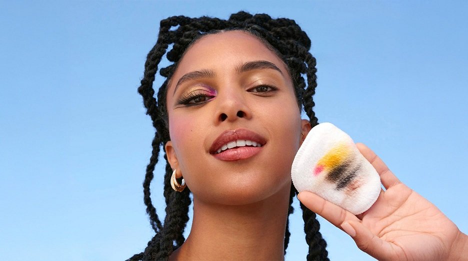 A woman with braids and colorful eye makeup holds a cotton pad showing removed makeup, demonstrating the effectiveness of micellar water.