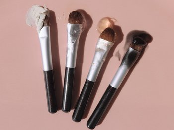 Several makeup brushes are shown with their bristles submerged in micellar water, demonstrating its use for cleaning makeup brushes.