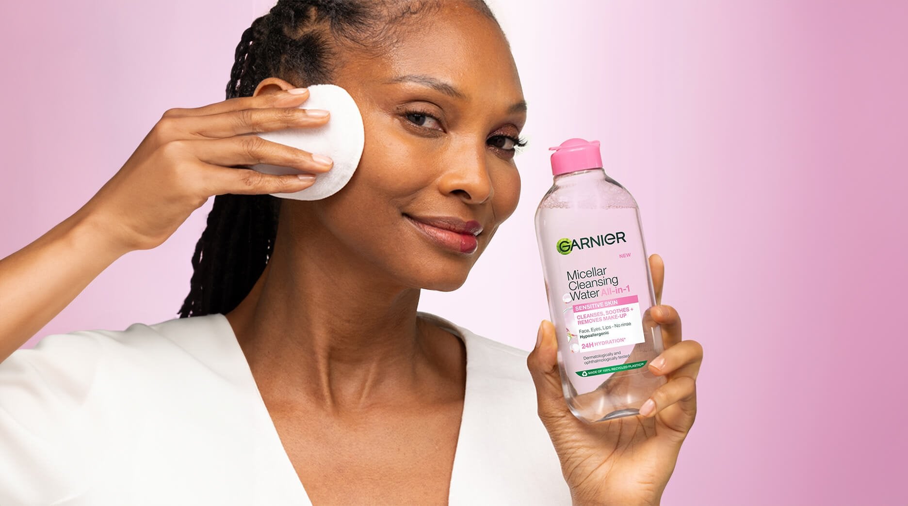 Garnier Micellar Cleansing Water Demonstration: A woman with braided hair smiling while using Garnier Micellar Cleansing Water All-in-1. She holds the large clear bottle with a pink cap in her left hand and uses a reusable white cleansing pad on her face with her right. The image features a "split-face" effect, showcasing the product's effectiveness by revealing clean, radiant skin on one half of her face while the other side remains made-up. The background is a soft pink gradient.