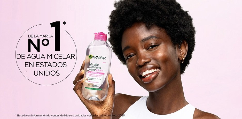 A smiling woman with short dark curly hair and deep skin tone holds up a clear bottle of Garnier Micellar Cleansing Water All-in-1 with a pink cap against a pink background, next to text reading "From the No. 1 Micellar Brand in America" with a footnote citing Nielsen sales data for Full Year 2025. 