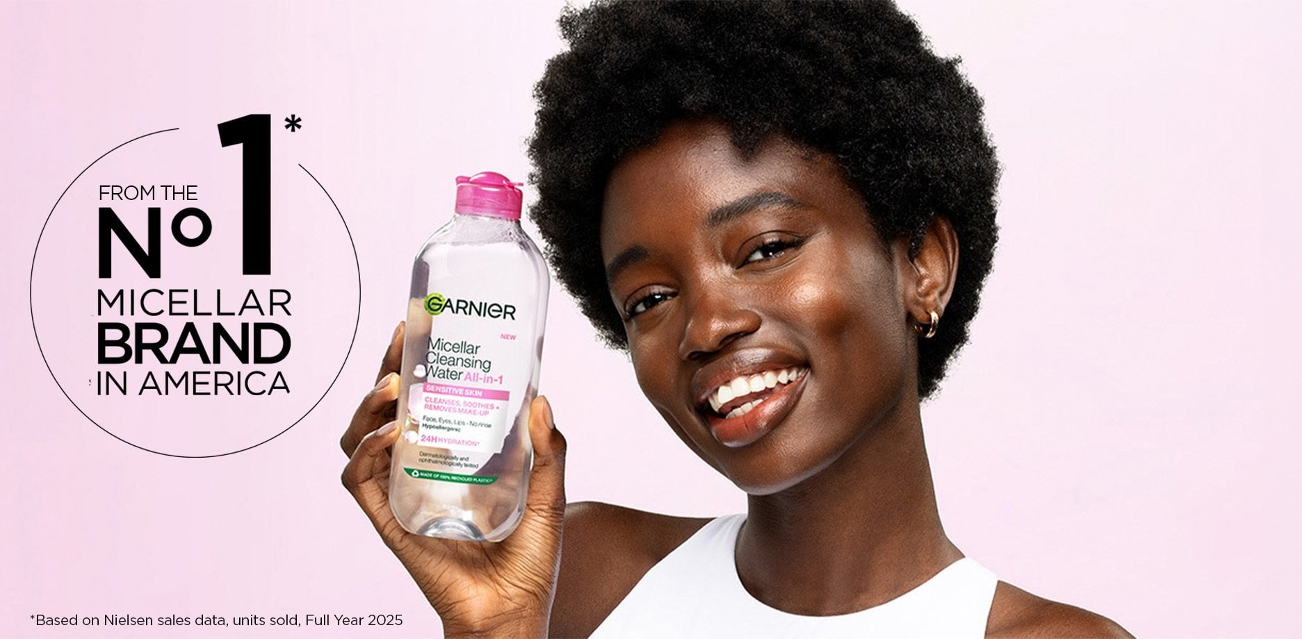 A smiling woman with short dark curly hair and deep skin tone holds up a clear bottle of Garnier Micellar Cleansing Water All-in-1 with a pink cap against a pink background, next to text reading "From the No. 1 Micellar Brand in America" with a footnote citing Nielsen sales data for Full Year 2025.