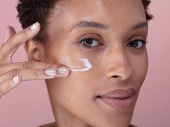 A person with dark skin applies a white cream moisturizer to their face.