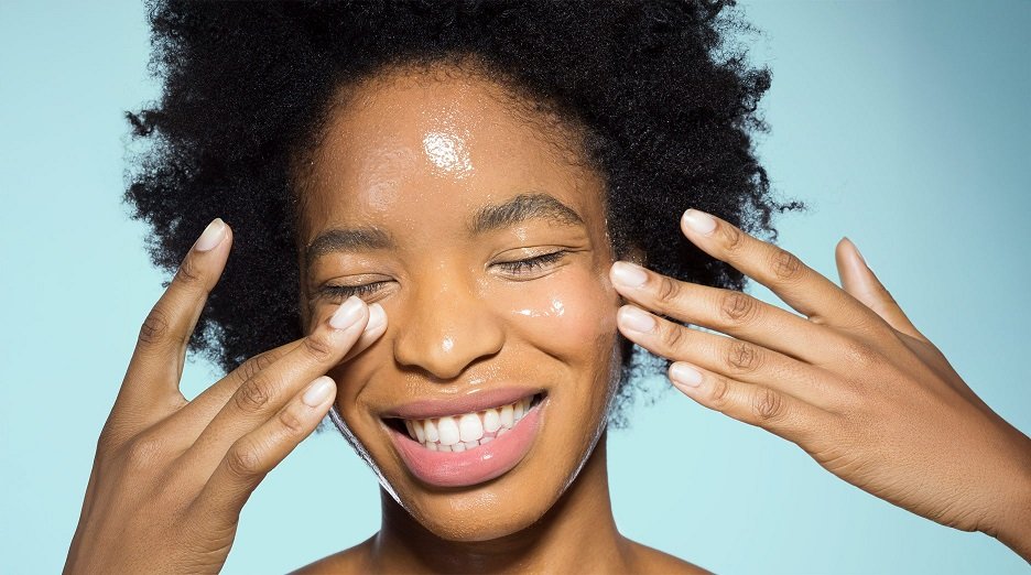 A person with dark skin applies a clear gel moisturizer to their face.