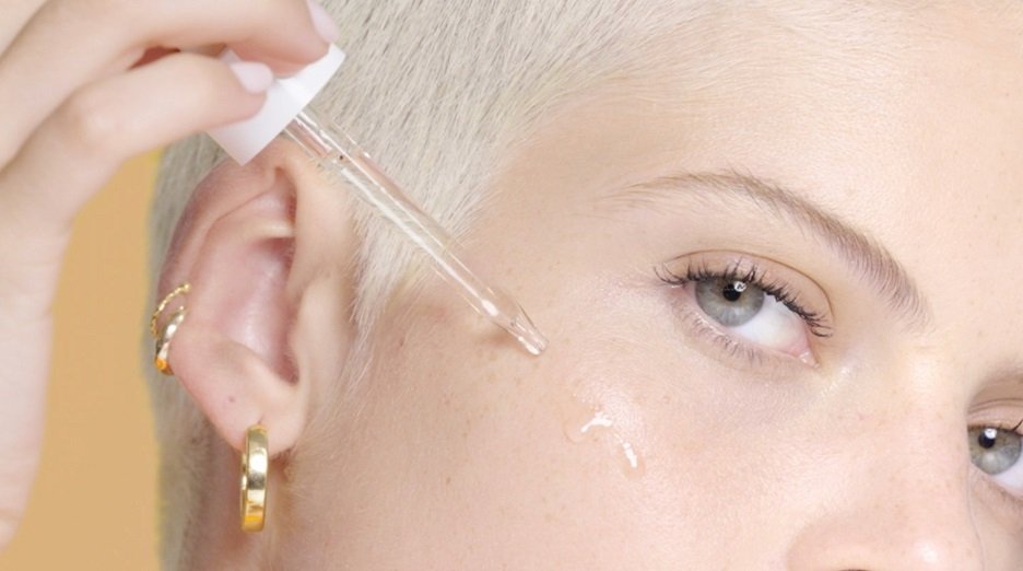 Model with Short Platinum pixie cut applying serum to side of her face.