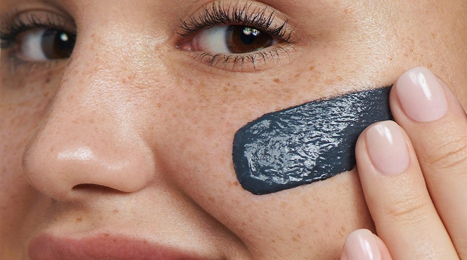 Close-up of a person applying a dark grey charcoal mask to their cheek.