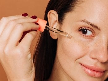 Close up of brunette woman looking in the mirror as she applies a clear serum across the top of her cheek.