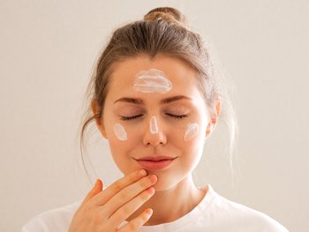 Front shot of blonde woman applying moisturizer to her clear skin. There are white streaks of moisturizer on her forehead, cheeks, and nose bridge. Her eyes are closed as she embraces the peace of her morning routine.