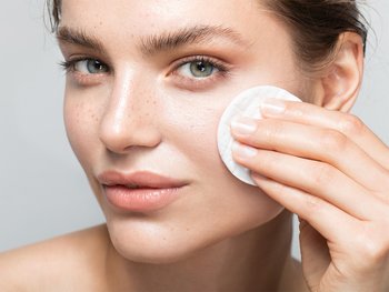 Close up front shot of woman with light skin and freckles using a cotton pad to apply toner to her cheek.