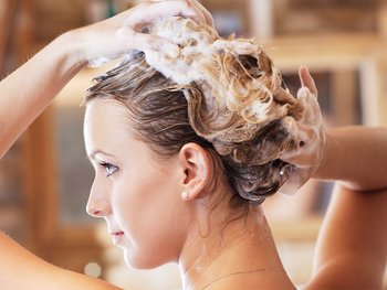Shot of top half of a woman washing her hair. We can see the backside of her from her shoulders up as her hair foams up with shampoo.