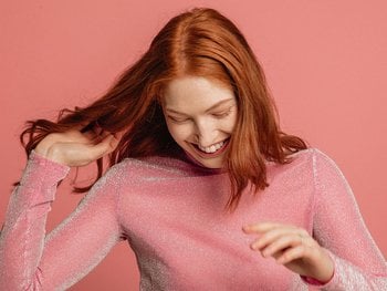 Front shot of smiling red-haired woman smiling as she looks down and dances. She looks chic in front of a pink-burgundy background.
