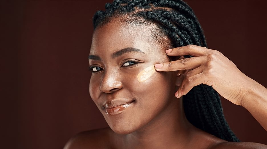 A woman with dark skin and braided hair applies BB cream or foundation to her cheek, demonstrating the application process.