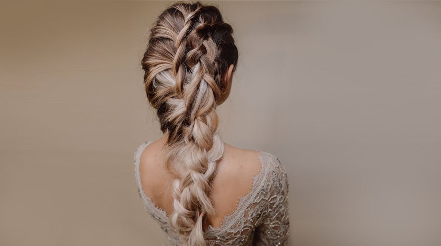 Girl with blonde balayage hair wears a chunky French braid, and stands against a beige background, facing away from the camera.
