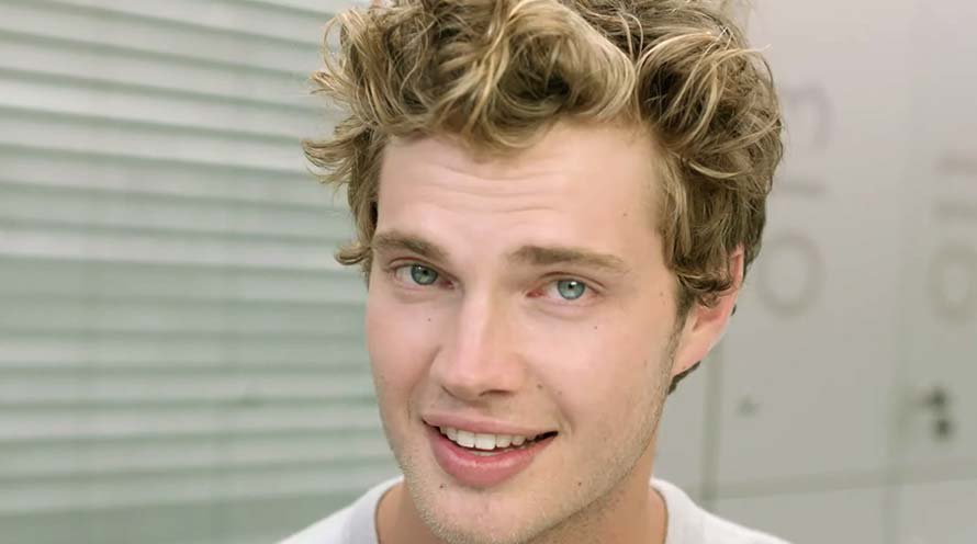 Shirtless man styles his curly light-brown hair with both hands, and stands against a white background.