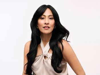 A woman with long, dark, wavy hair, wearing a light-colored halter-neck top, looking confidently at the camera against a white background.