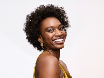 A smiling woman with short, brown, natural curly hair and a gold earring, looking over her shoulder at the camera, wearing a yellow top against a white background.