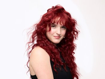 A woman with long, vibrant red, curly hair and bangs, smiling subtly and looking slightly to the side, wearing a black top against a white background.
