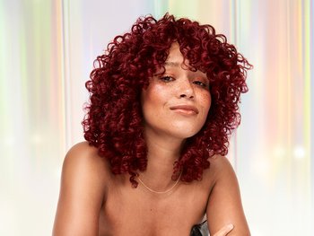 A woman with vibrant reddish-brown curly hair and freckles, looking directly at the camera with a subtle smile, against a shimmering, iridescent background.