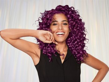 A joyful woman with long, vibrant purple curly hair, smiling broadly and posing with one hand behind her head, against a solid black background. She is wearing a black sleeveless top.