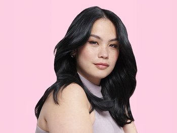 A young woman with shoulder-length, dark, wavy hair parted to the side, looking confidently at the camera against a pink background. She has freckles and subtle makeup. She has applied Color Retouch Black to her roots.