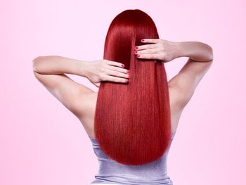 The back of a person with very long, straight, vibrant red hair, reaching down their back. Their hands are gently placed on the sides of their hair, showing off red nail polish, against a pink background. She has applied Color Retouch Red to her roots.