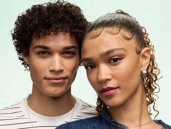 A young man with short brown curls and a young woman with light brown curly hair pulled back, both showcasing defined, healthy curl texture achieved with Garnier Fructis Curl Styling Products.
