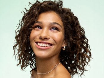A joyful woman with shoulder-length, deeply defined brown curls smiling upward against a light background. Defined curls obtained using Garnier Fructis Curl Styling products.