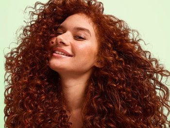 A smiling woman with voluminous, deep auburn coily curls cascading past her shoulders against a soft green background. She showcases the volume and definition possible with Garnier Fructis Curl Styling Products.